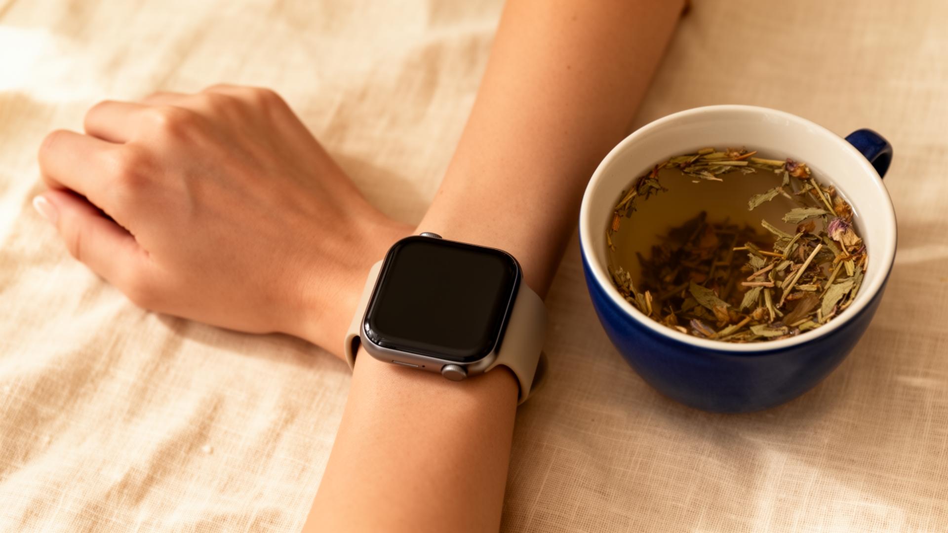 A woman's wrist wearing a smartwatch beside a cup of herbal tea on warm linen — a calm morning wellness scene